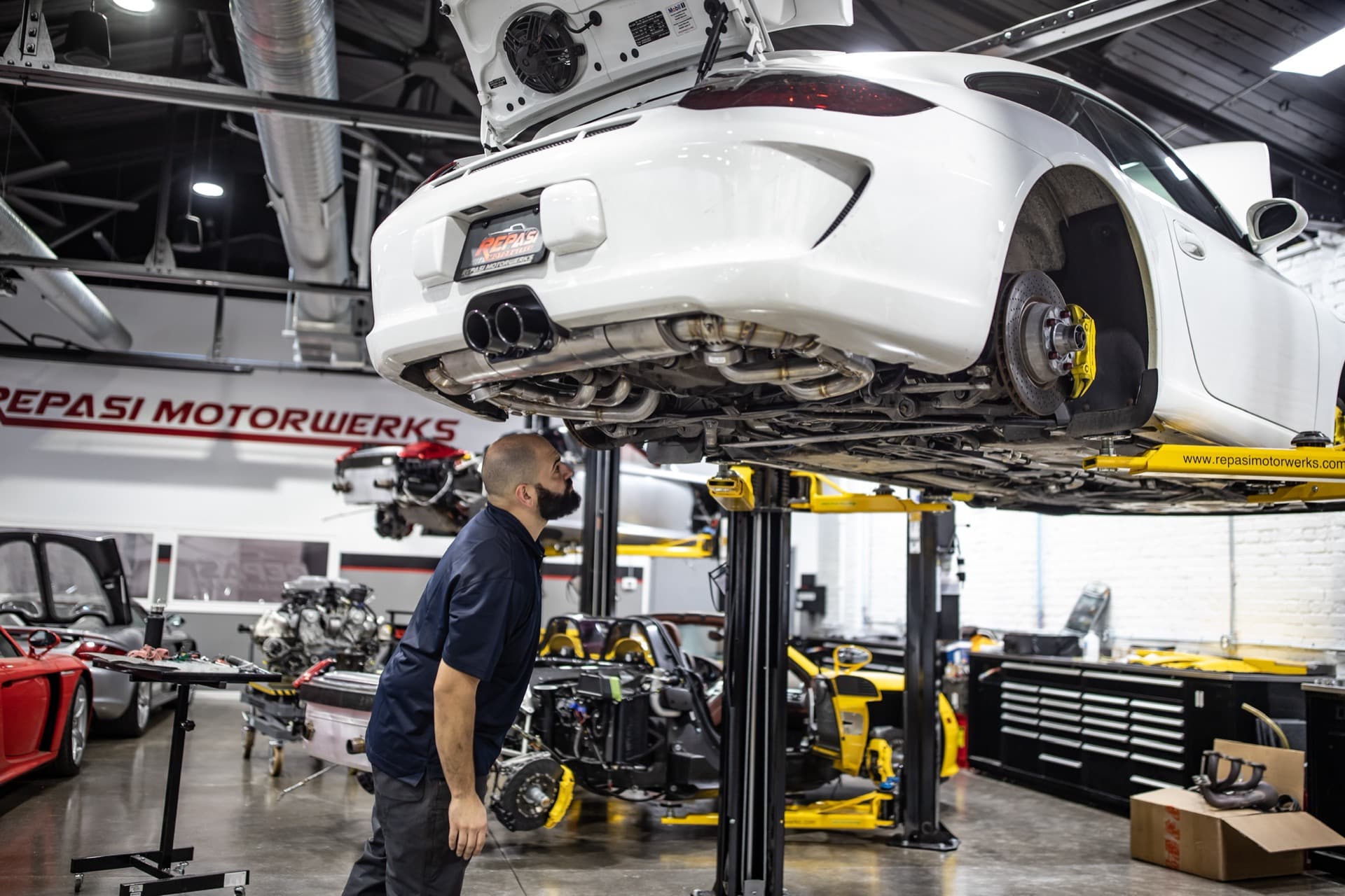 Technician inspecting the underside of a white Porsche 991 GT3 on a lift during a pre-purchase inspection at Repasi Motorwerks